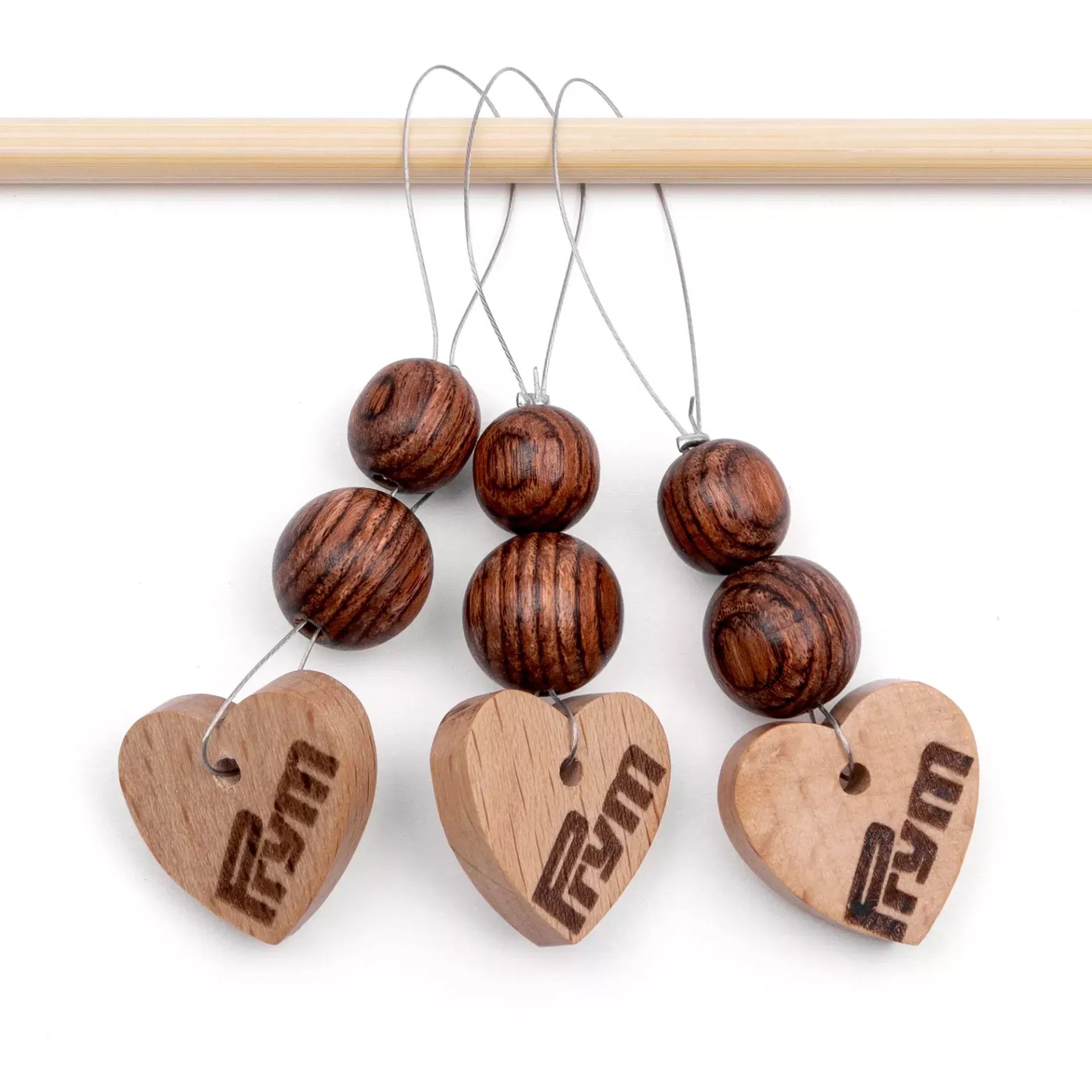 Three heart-shaped wooden stitch markers with dark beads hanging from a light wood knitting needle.