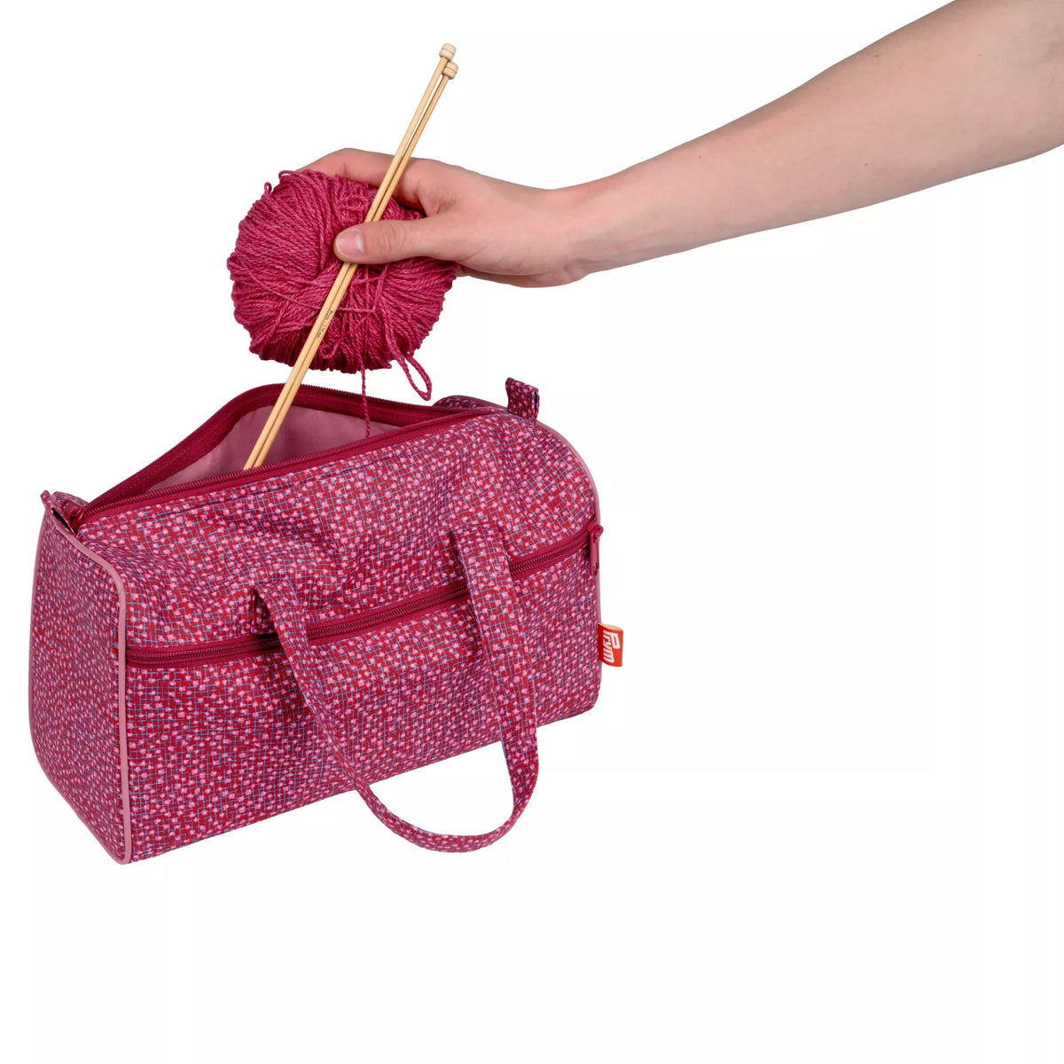 Burgundy project bag with a ball of pink yarn and knitting needles being placed inside.