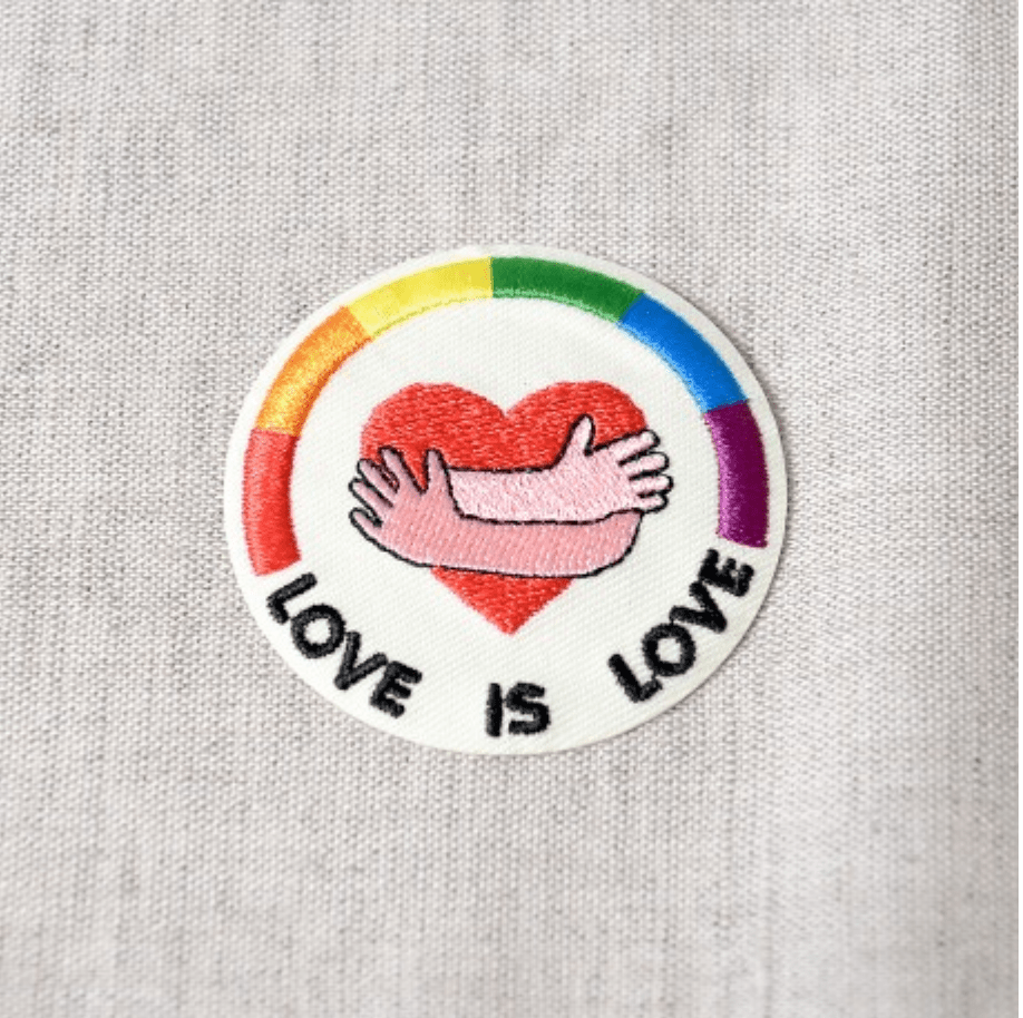 A circular patch with a rainbow border, a red heart with pink arms hugging it, and the words "LOVE IS LOVE".