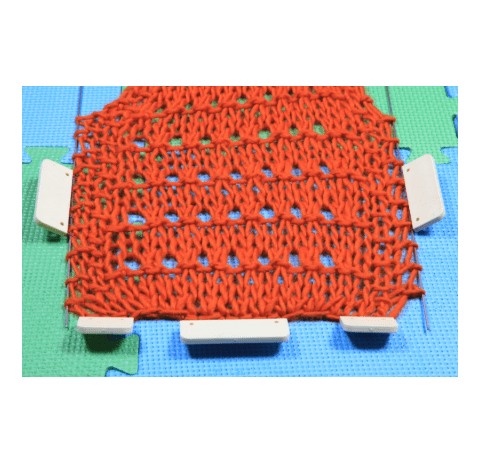 Orange knitted fabric stretched on blue and green puzzle mats with white clips.