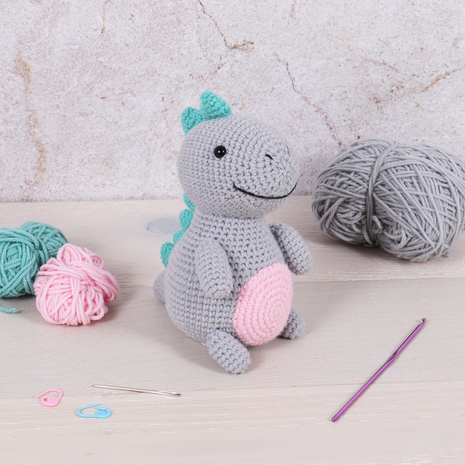 A completed grey crochet dinosaur amigurumi sits on a table with yarn, a hook, and stitch markers.