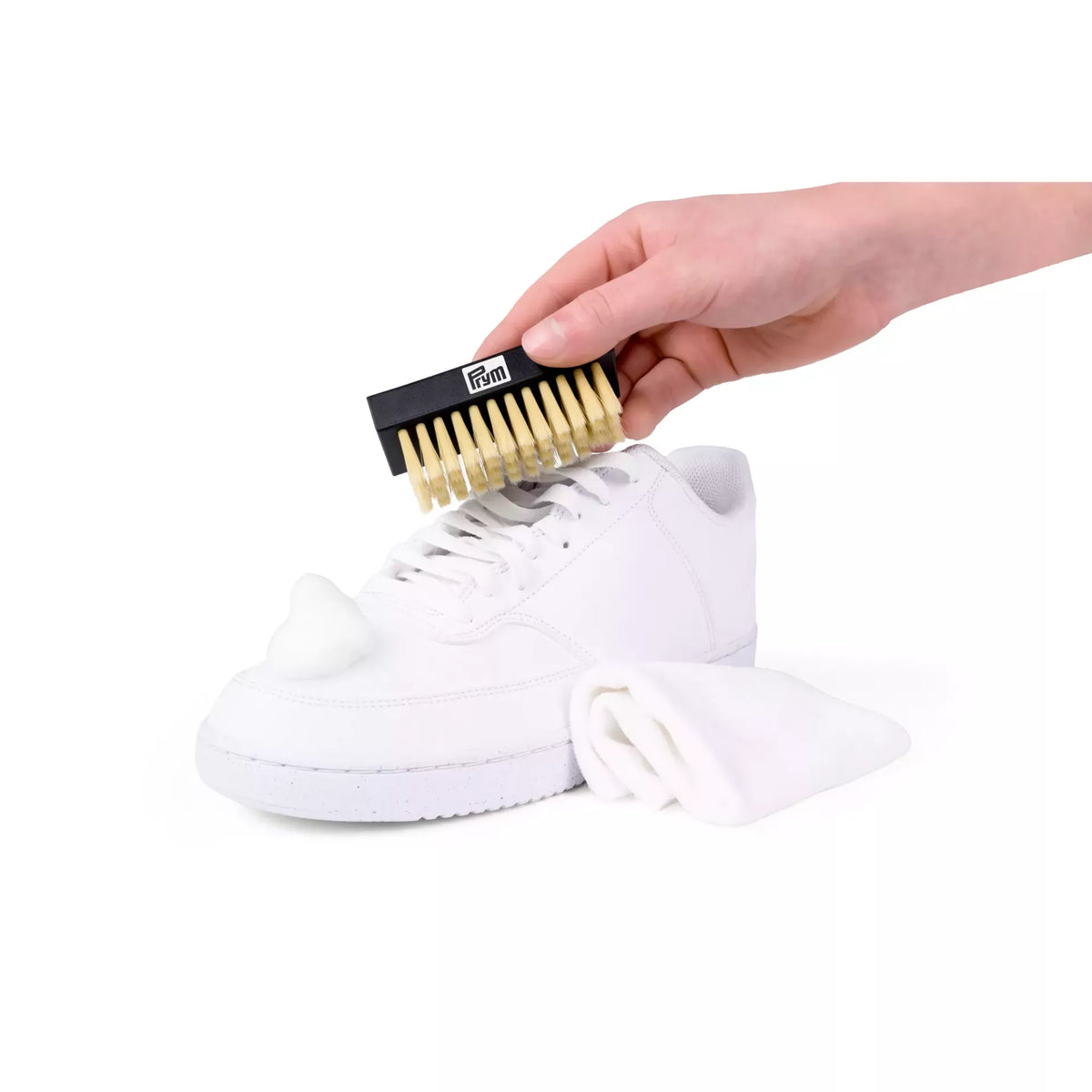 Sneaker care set being used to clean a white shoe with foam, a brush, and a microfiber cloth.
