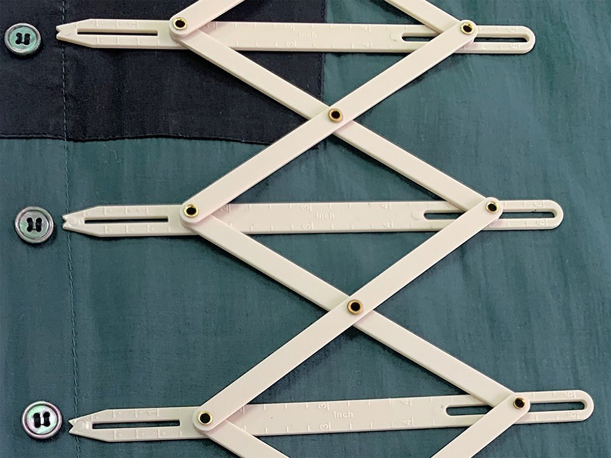 Expandable white plastic Hemline sewing gauge measuring distances between buttons on green fabric.