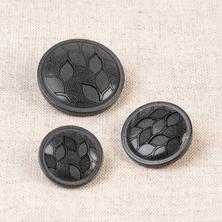 Three black polyester shank buttons with an embossed leaf design in varying sizes on a beige fabric.