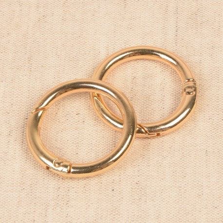 Two gold-colored round metal spring gate clips for bags resting on a neutral fabric background.