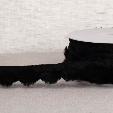 Black synthetic fur pompom trim on a spool against a neutral fabric background.
